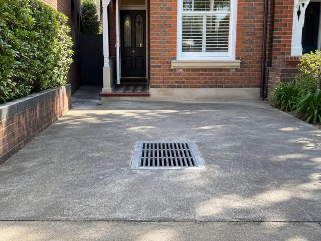 Clean driveway drain grate beside a brick house and garden beds in Woollahra